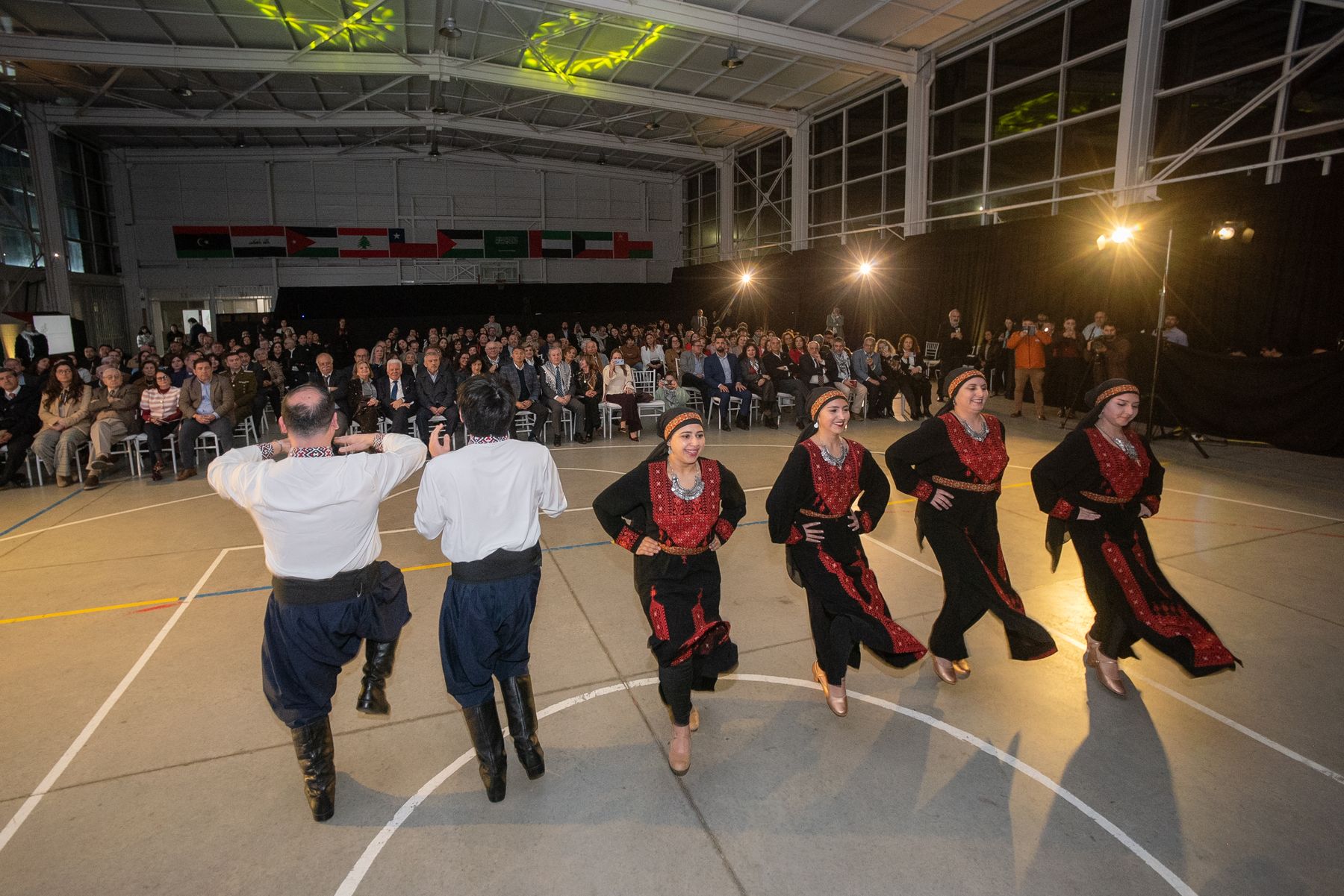 Bailarinas con vestidos negros bordados en rojo y bailarines con camisa blanca danzan frente al público